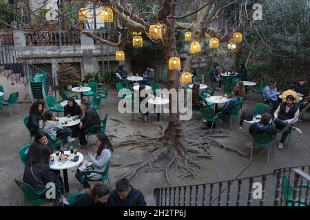 Barcellona, Spagna - 20 settembre 2021, lanterne multicolore sono appese sugli alberi e brillano al crepuscolo. La gente si siede ad un tavolo nel giardino ed ha dinne Foto Stock