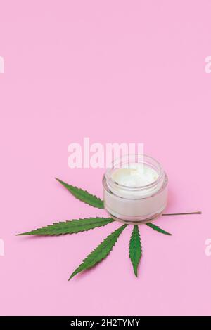 Vaso in vetro cosmetico con crema facciale infusa di cannabis e foglia verde di marijuana su sfondo rosa pastello, immagine verticale Foto Stock