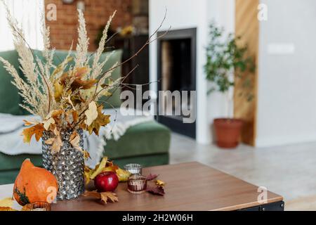 Interni decorati in autunno. Bouquet di fiori gialli in vaso di vetro su elegante tavolo di legno, zucca, mele rosse, foglie, candele, coperta. Grana Foto Stock