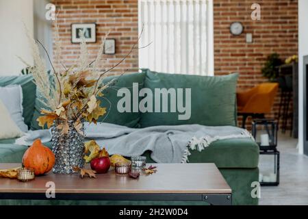 Interni decorati in autunno. Bouquet di fiori gialli in vaso di vetro su elegante tavolo di legno, zucca, mele rosse, foglie, candele, coperta. Grana Foto Stock
