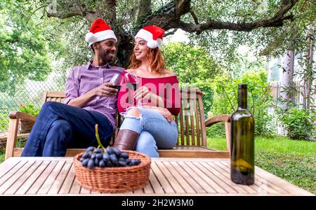 buon natale! giovane coppia di persone diverse che festeggia il natale indossando un cappello di santa godendo il vino all'aperto in un giardino. amici che fanno un brindisi celebratoty Foto Stock