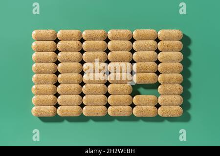 Pillole multivitaminiche marroni disposte in ordine rettangolo, una pillola prelevata su sfondo verde. Compresse di erbe giornaliere disposte nella forma quadrata. Foto Stock