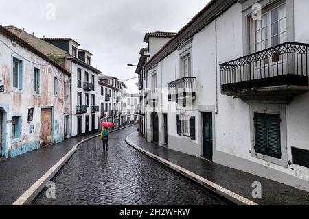 Donna in gialle giacca da pioggia e ombrello rosso che cammina su strada vuota in una giornata piovosa a Ponta Delgada, Sao Miguel, Azzorre, Portogallo Foto Stock