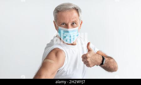 Vaccinato Senior Man gesturing Thumbs-Up su sfondo grigio, indossare maschera Foto Stock