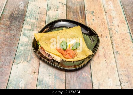 Crepe salata con salmone norvegese, uova sode, foglie di basilico e piatto nero su tavola di legno Foto Stock