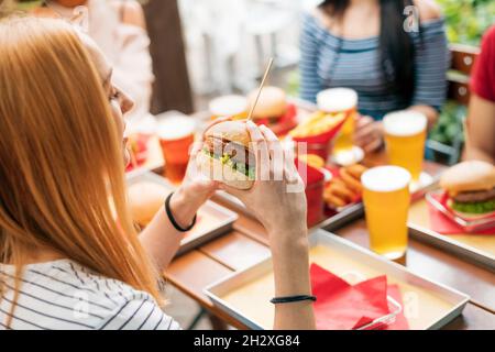 Vista laterale della giovane donna che si prepara a bocciare l'hamburger gustoso intero durante il pranzo con gli amici al tavolo con spuntini e bevande nel ristorante Foto Stock