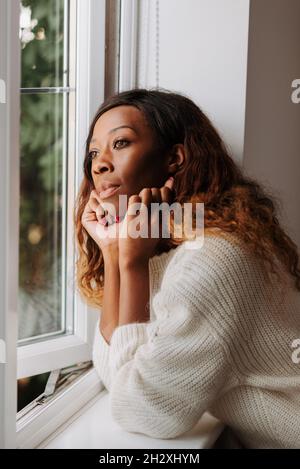 Una bella giovane donna nera elegante appoggiata sul davanzale che guarda fuori dalla finestra Foto Stock