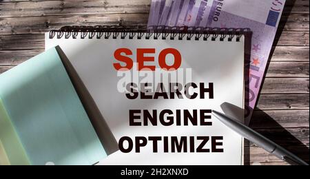 SEO SERACH MOTORE OTTIMIZZARE parola di testo abbreviazione scritta in blocco note sul tavolo da ufficio, accanto alle banconote in euro. Concetto di business KPI Foto Stock