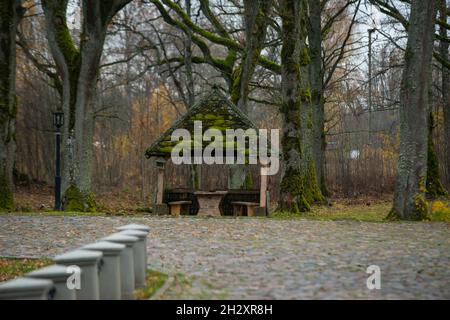 vecchio gazebo in legno con un tetto sovrastato di muschio verde nel parco con un parcheggio lastricato sotto vecchi querce in una giornata di sole autunno Foto Stock