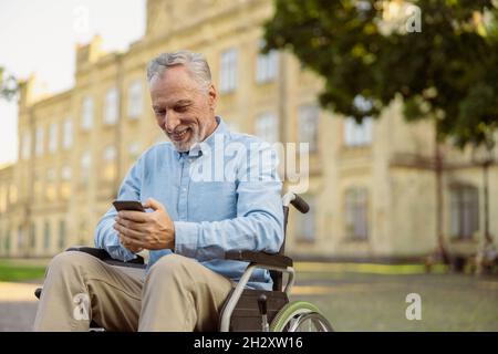 Uomo anziano allegro, che recupera il paziente maschile in sedia a rotelle utilizzando lo smartphone mentre si riposa da solo nel parco vicino all'ospedale Foto Stock