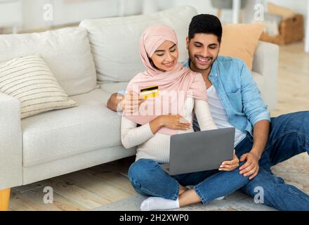 Amorevole famiglia araba in attesa di utilizzare il portatile e la carta di credito, shopping online, seduta sul pavimento in soggiorno interno Foto Stock