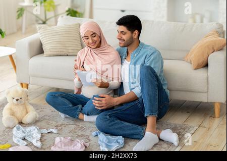 Amorevole coppia musulmana incinta che si prepara per il parto, facendo checklist mentre si siede con i vestiti del bambino intorno Foto Stock