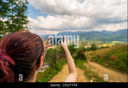 Vista posteriore della donna che tiene lo smartphone in entrambe le mani durante l'escursione, scattare foto per blog di viaggio, cercare un nuovo percorso online, cercare la connessione Foto Stock