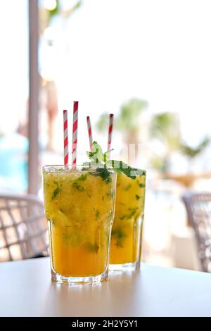 Primo piano su due bicchieri di mojito alcolico al mango arancione o bevande analcoliche con cannuccie su tavolo in terrazza estiva, nessuna persona Foto Stock