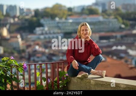 Una donna in blusa con cappuccio, seduta all'aperto contro lo sfondo sfocato della città. Foto Stock