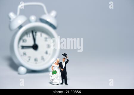 Orario del matrimonio e programma concetto persone in miniatura, giocattoli fotografia. Sposi e sposi in piedi davanti all'orologio isolato su sfondo bianco. Immagine p Foto Stock