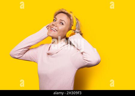 Donna caucasica bionda dai capelli corti che ascolta musica. Ritratto su sfondo giallo. Sorridendo e guardando la fotocamera che tiene le mani fino a lui Foto Stock