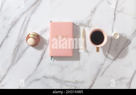 Diario rosa di colore corallo per l'anno 2022, penna, caffè, biscotti di macaron. Sfondo in marmo Foto Stock