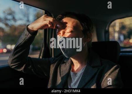 Donna d'affari con grave mal di testa sintomi di dolore e maschera protettiva al sedile posteriore di un'auto, fuoco selettivo Foto Stock