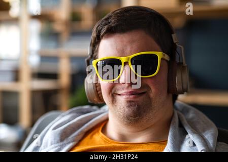 Primo piano del giovane felice con sindrome di Down con occhiali da sole e cuffie seduti in interni Foto Stock