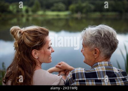 Vista posteriore della madre anziana felice che tiene le mani con la figlia adulta quando si siede vicino al lago all'aperto in natura. Foto Stock