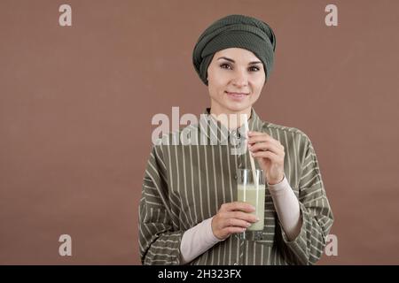Giovane donna sorridente con un bicchiere di cocktail al latte o un frullato davanti alla fotocamera su sfondo marrone Foto Stock