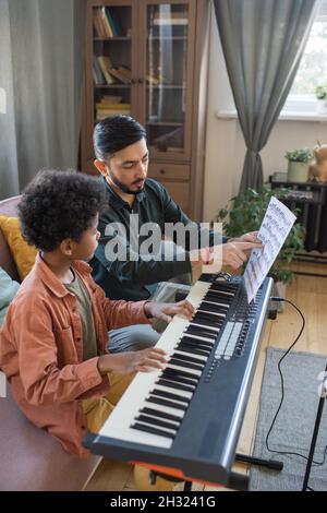 Insegnante sicuro di musica che indica le note sulla carta mentre insegna a ragazzo piccolo carino come suonare la tastiera del pianoforte Foto Stock