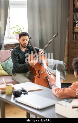 Giovane insegnante di musica fiduciosa che spiega al ragazzo come suonare il violoncello mentre si siede sul divano davanti alla sua pupilla Foto Stock