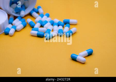 Due capsule blu-bianche pillole su blur molti di pillole versando da bottiglia di droga di plastica. Capsule su sfondo giallo. Farmaci da prescrizione. Farmacia Foto Stock