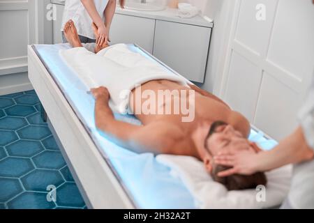 L'uomo rilassato si trova sul divano luminoso mentre i terapisti fanno massaggi nel salone spa Foto Stock