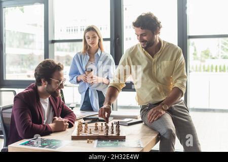 Uomini d'affari sorridenti che giocano a scacchi vicino a un collega sfocato con il caffè in ufficio Foto Stock