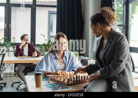 Donne d'affari interrazziali che giocano a scacchi vicino a un collega offuscato in ufficio Foto Stock