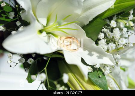 Anelli di nozze che posano su bouquet nuziale con giglio bianco. Primo piano sugli accessori da sposa. Classici bands dorati per matrimoni. Foto Stock