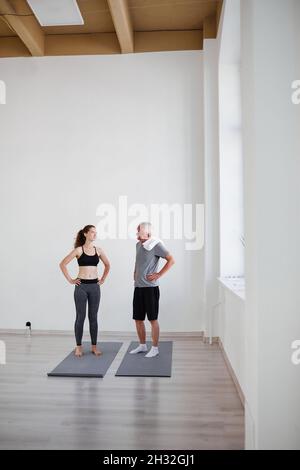 Uomo anziano che parla con il suo allenatore mentre si alza sui tappetini durante l'addestramento personale in studio Foto Stock