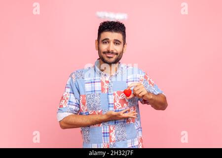 Ritratto di attraente uomo bearded con nimb sopra la testa che tiene il piccolo cuore rosso nelle mani, guardando la macchina fotografica con amore, indossando la camicia casual blu. Studio interno girato isolato su sfondo rosa. Foto Stock