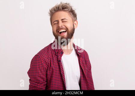 Ritratto di divertente bel ragazzo portato in camicia a quadri in piedi con occhi chiusi e ridendo. Studio interno girato isolato su sfondo grigio Foto Stock