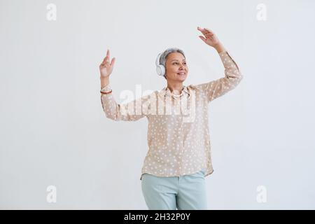 Donna anziana felice in cuffie wireless che ascoltano musica e ballano sullo sfondo bianco Foto Stock