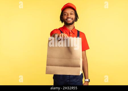 Ritratto di positivo sorridente deliveryman in tute che danno ordinato pacco, consegna cibo in sacchetto di carta, servizi postali. Studio interno girato isolato su sfondo giallo. Foto Stock