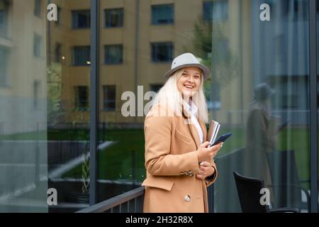 donna in un cappotto e cappello beige che tiene un notebook e uno smartphone Foto Stock