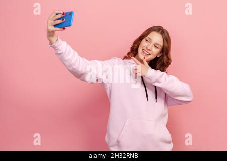 Ritratto di sorridente ragazza teen con capelli ricci in felpa con cappuccio prendendo selfie, avendo conversazione sulla videochiamata o in streaming blog, puntando il dito. Studio interno girato isolato su sfondo rosa Foto Stock