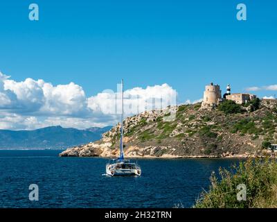 Catamarano in mare e torre faro vicino albero sempreverde lungo la riva fiancheggiata da montagne di Calamosca in Sardegna Italia in una giornata di sole, con sk blu Foto Stock