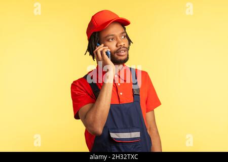 Ritratto di corriere o di riparatore in tute prendendo telefono con il cliente, specificare l'ora e il luogo di completamento del lavoro o la consegna della merce. Studio interno girato isolato su sfondo giallo. Foto Stock