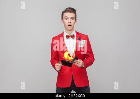 Ritratto di giovane mago sorpreso con capelli alla moda in tuxedo rosso e bow tie tenere portafoglio sul fuoco, guardando scioccato e spaventato da borsa bruciante. Studio interno girato isolato su sfondo grigio Foto Stock