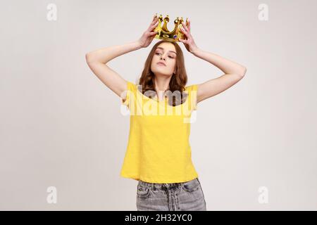 Ritratto di egoista ragazza teenage capelli castani in giallo T-shirt che tiene la corona d'oro sopra la testa, avendo espressione di arroganza, stato privilegiato. Studio interno girato isolato su sfondo grigio. Foto Stock