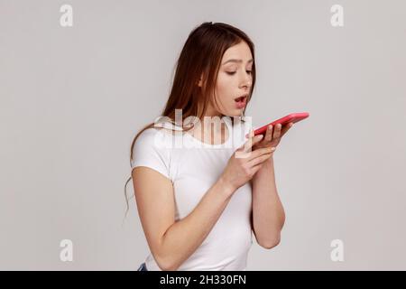 Donna attraente che tiene il telefono cellulare con entrambe le mani e parla in altoparlante di carico, facendo nota vocale, assistente vocale, indossando una T-shirt bianca. Studio interno girato isolato su sfondo grigio. Foto Stock