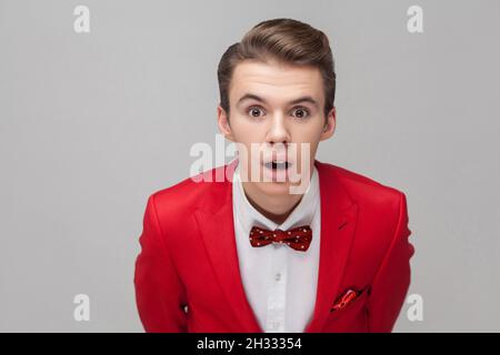 Ritratto di bel Signore con capelli eleganti in tuxedo rosso e bow tie in piedi con occhi grandi e bocca aperta, guardando la fotocamera in stupore, shock. studio girato isolato su sfondo grigio Foto Stock