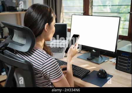 Ragazza asiatica freelance che usa il telefono cellulare al tavolo di lavoro con monitor vuoto in casa Foto Stock