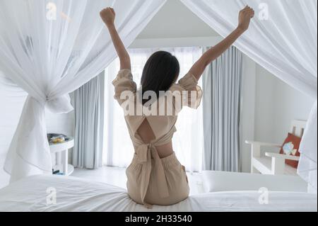 Retro della donna asiatica che si allunga dopo essersi svegliati in camera bianca con zanzariera al mattino Foto Stock