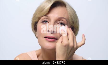 donna di mezza età che applica fondazione faccia con puff cosmetico isolato su bianco Foto Stock