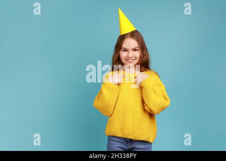 Bambina in cono di festa che guarda sorridendo alla macchina fotografica e che punta a se stessa, festeggia il compleanno, indossando il maglione giallo casual stile. Studio interno girato isolato su sfondo blu. Foto Stock